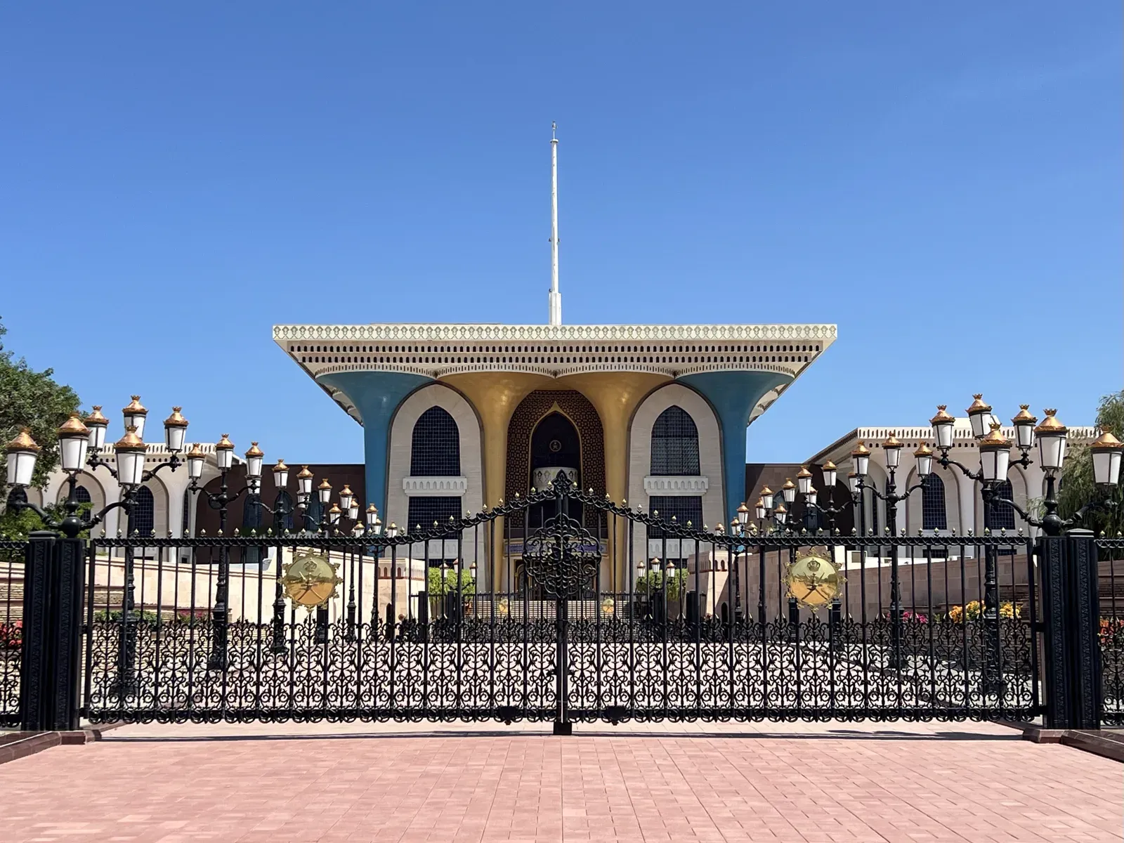 Het Al Alam Palace in oud Muscat achter het sierlijke smeedijzeren hek, met de blauw-gouden gevel en lantaarns onder een strakblauwe lucht