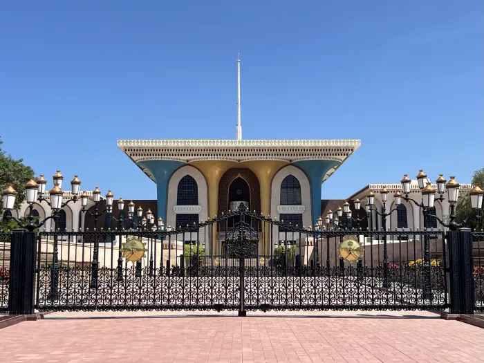 Het Al Alam Palace in oud Muscat achter het sierlijke smeedijzeren hek, met de blauw-gouden gevel en lantaarns onder een strakblauwe lucht