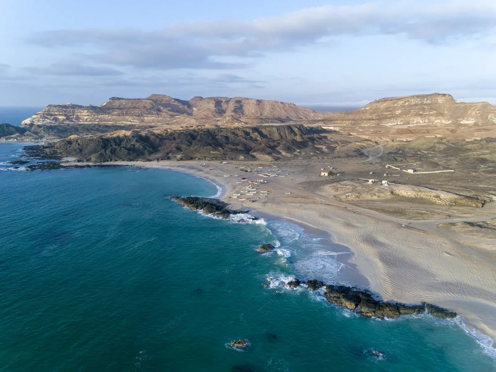 Luchtfoto van het brede strand van Al Duqm met turquoise water, rotspunten in zee en getrapte kliffen op de achtergrond