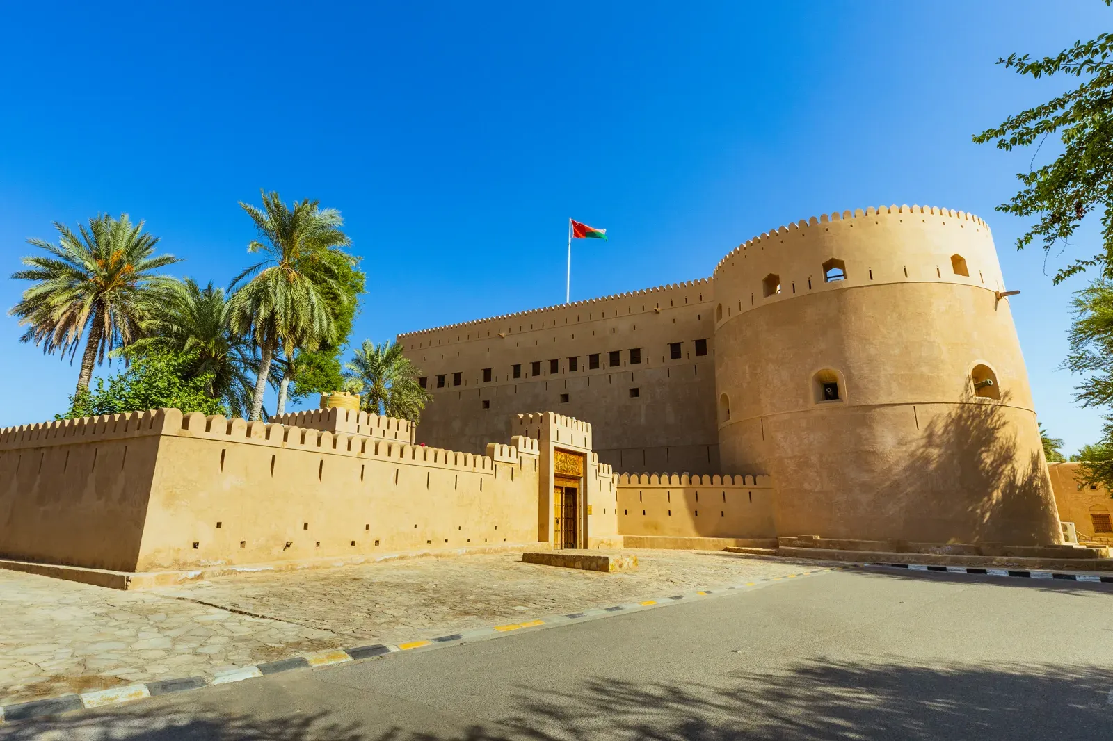 Het massieve Al Hazm-fort bij Rustaq met zijn ronde toren en de Omaanse vlag, achter een lemen vestingmuur tussen de palmen