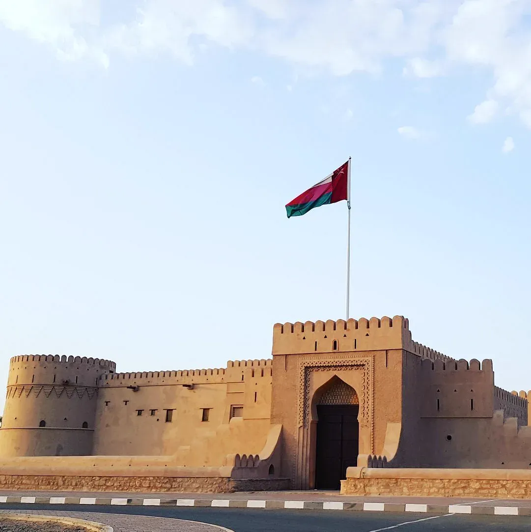 Het gerestaureerde Al Khandaq Fort met ronde hoektoren, versierde poort, gekanteelde muren en de Omaanse vlag