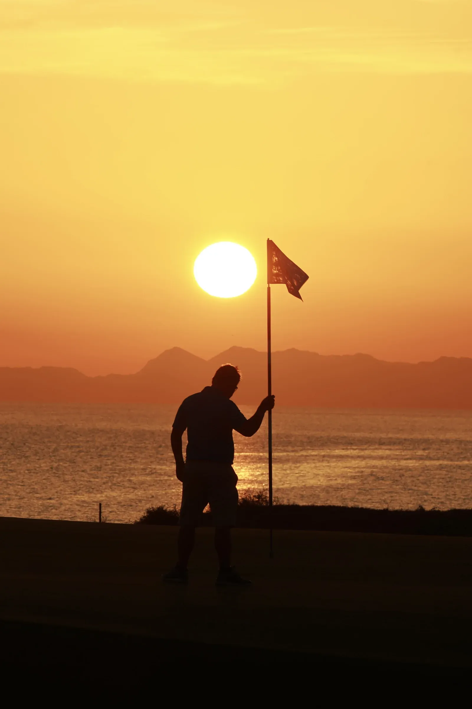 Een golfer als silhouet bij de vlag op Al Mouj Golf tijdens een gouden zonsondergang boven de bergen