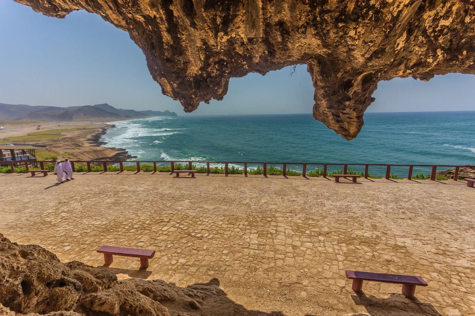 Uitzicht vanuit de Al Marnif-grot bij Al Mughsail over een geplaveid terras met bankjes en de kustlijn van Dhofar