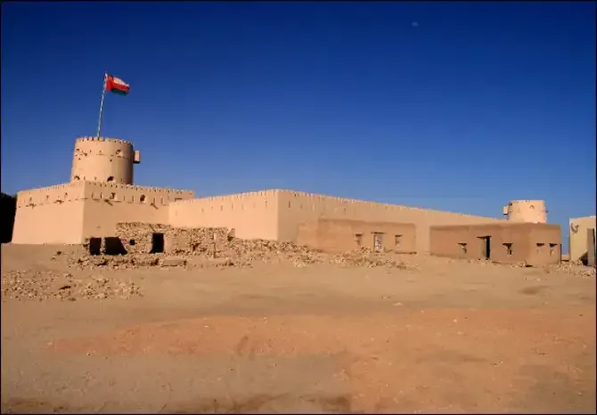 Al Sunaysilah Fort bij Sur met de Omaanse vlag op de ronde hoektoren, gezien vanaf het zandvlak onder een diepblauwe lucht
