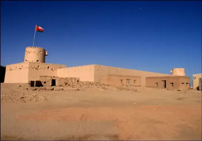 Al Sunaysilah Fort bij Sur met de Omaanse vlag op de ronde hoektoren, gezien vanaf het zandvlak onder een diepblauwe lucht