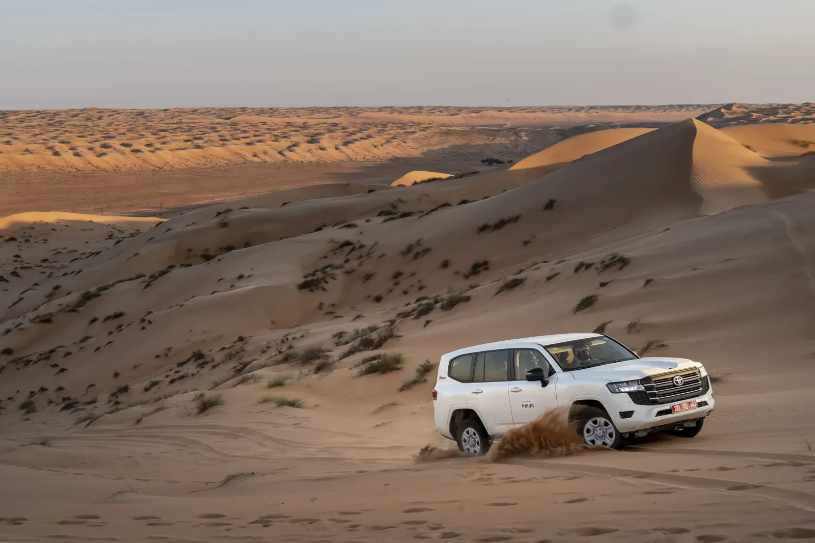 Een witte terreinwagen rijdt over de zachtgolvende duinen van de Wahiba Sands in het warme avondlicht