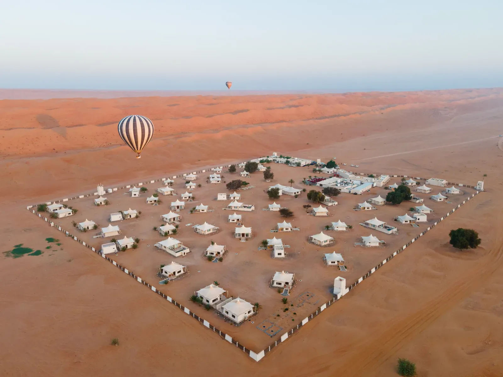 Luchtfoto van een ommuurd woestijnkamp met tenten en lodges tussen de duinen van de Wahiba Sands, met een luchtballon erboven