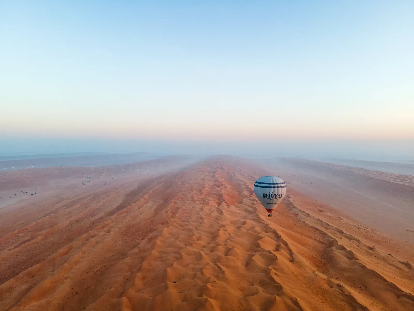 Een luchtballon zweeft bij dageraad laag over de eindeloze, roodgekleurde duinkammen van de Wahiba Sands