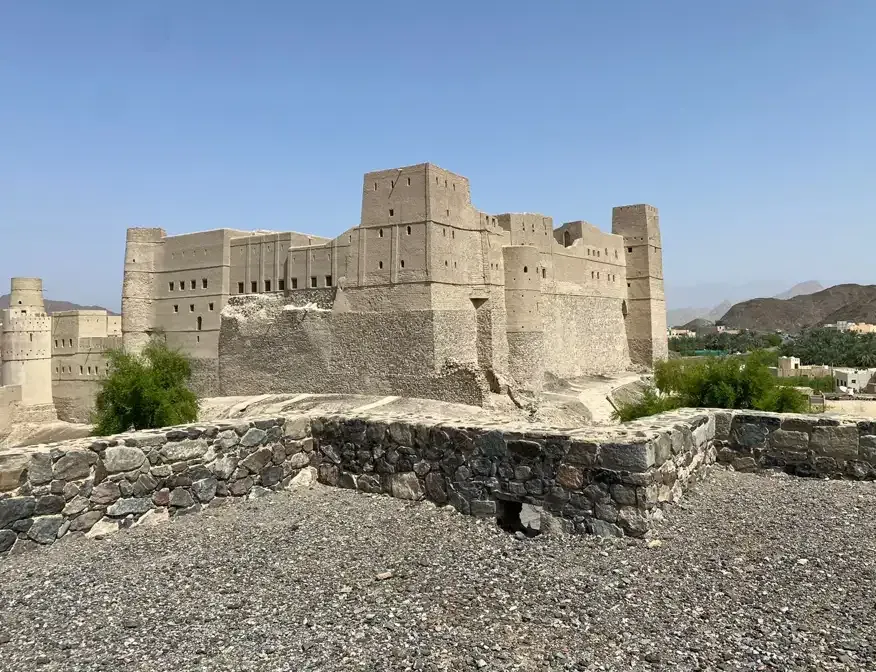 De robuuste lemen muren en torens van het Bahla-fort gezien vanaf een stenen wal, met de Hajar-bergen op de achtergrond