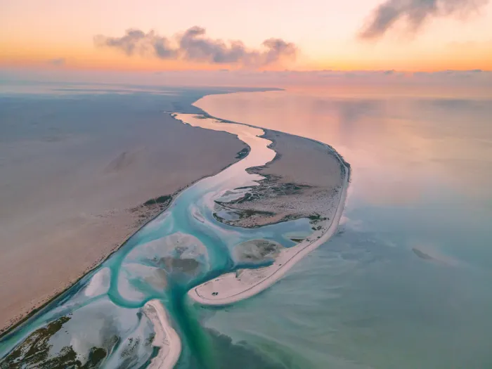 Luchtfoto bij zonsondergang van de getijdenvlakten en lagunes van Bar Al Hikman, met slingerende geulen in roze en turquoise tinten
