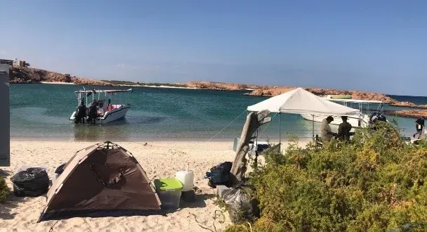 Een tentje en parasol op het witte strand van een Daymaniat-eiland, met een boot in de turquoise baai