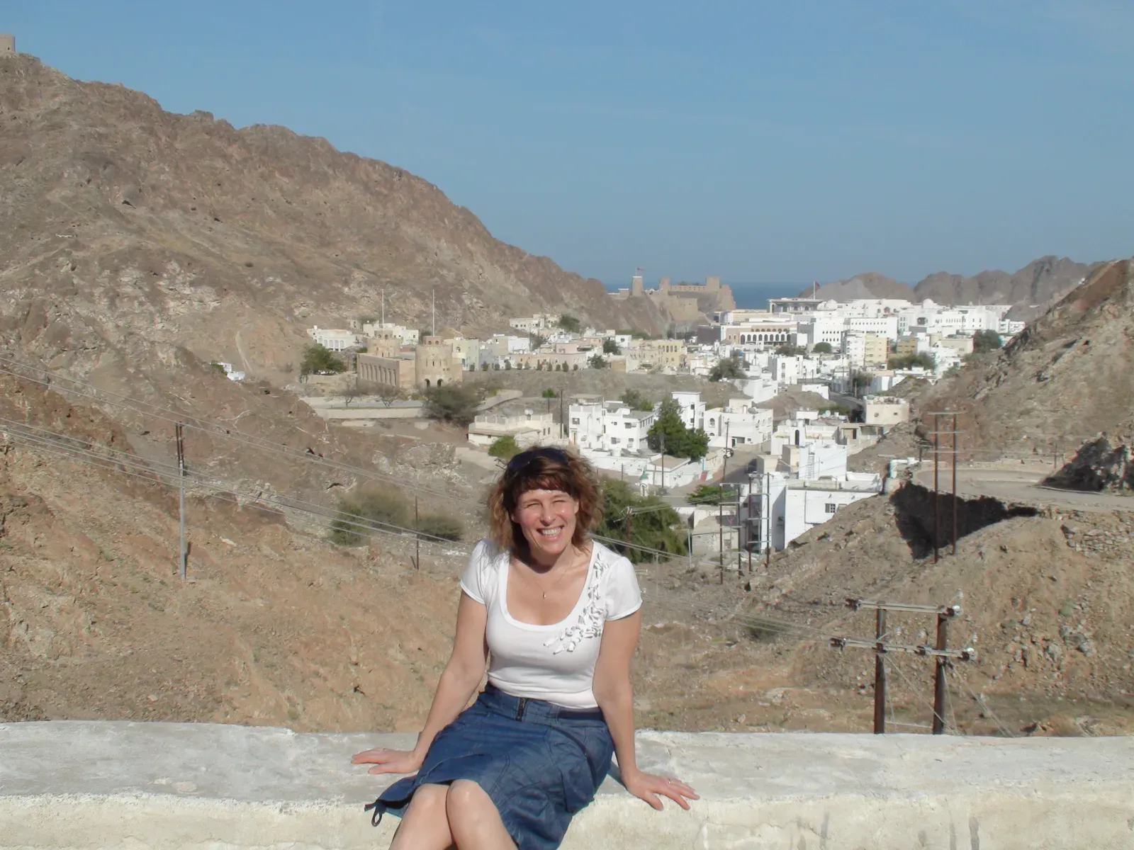 Danielle zit op een muurtje met uitzicht over Oud-Muscat, met het fort en de witte huizen tussen de bergen en de zee
