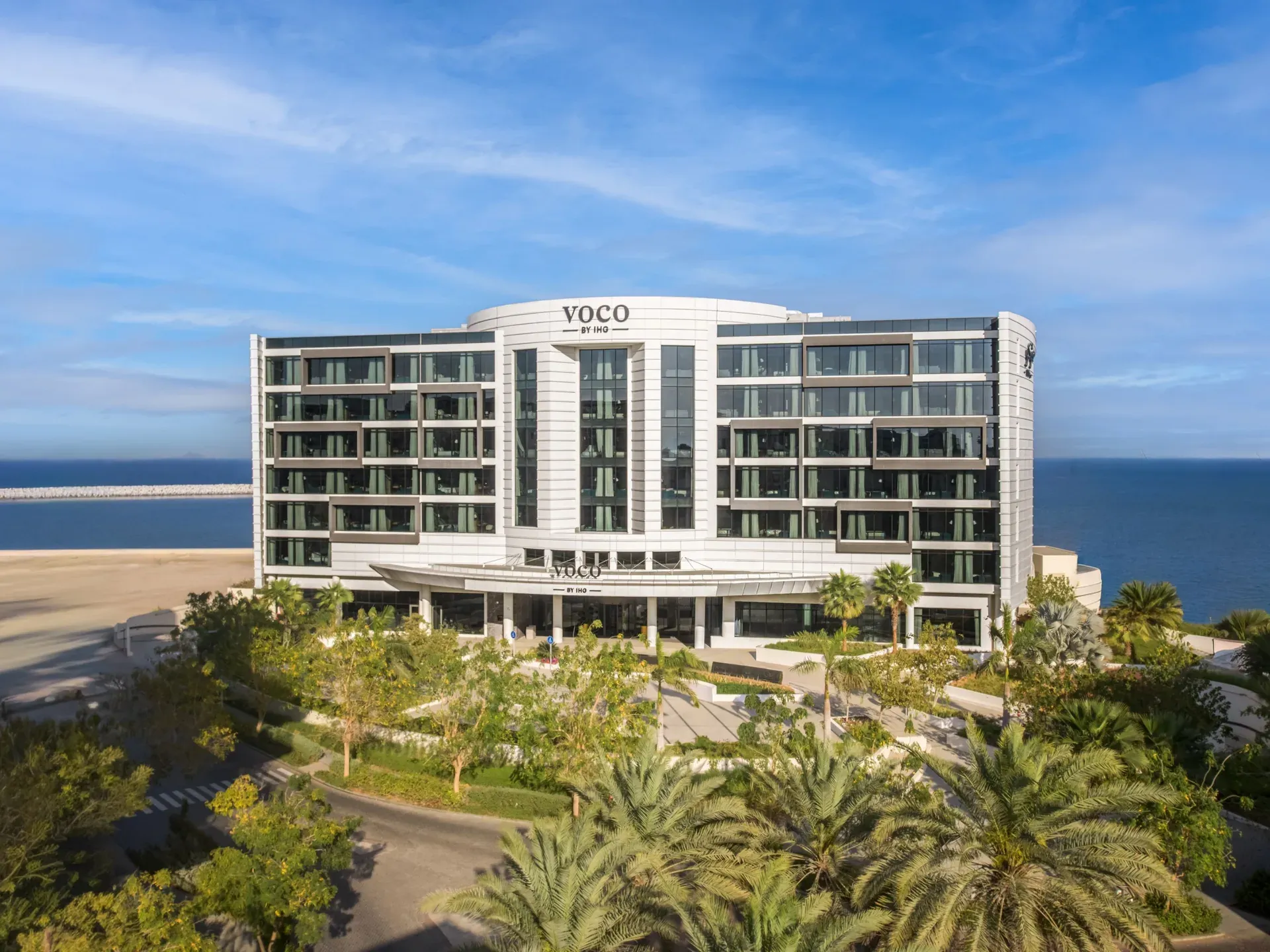 Het voco Muscat Al Mouj, een modern hotelgebouw aan zee met palmen en tuinen ervoor