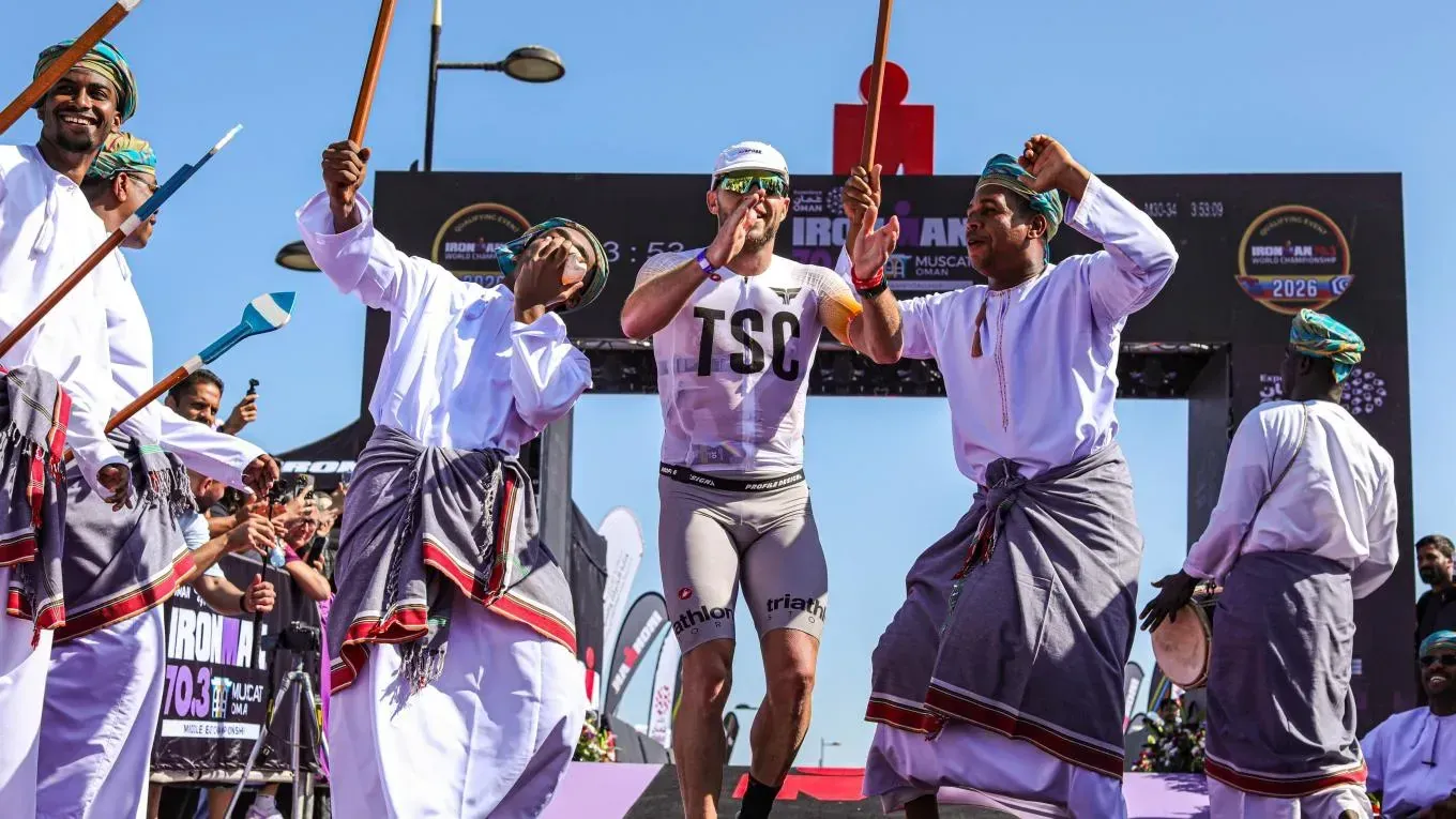 Een triatleet finisht bij IRONMAN 70.3 Muscat, juichend onthaald door traditionele Omaanse dansers met stokken