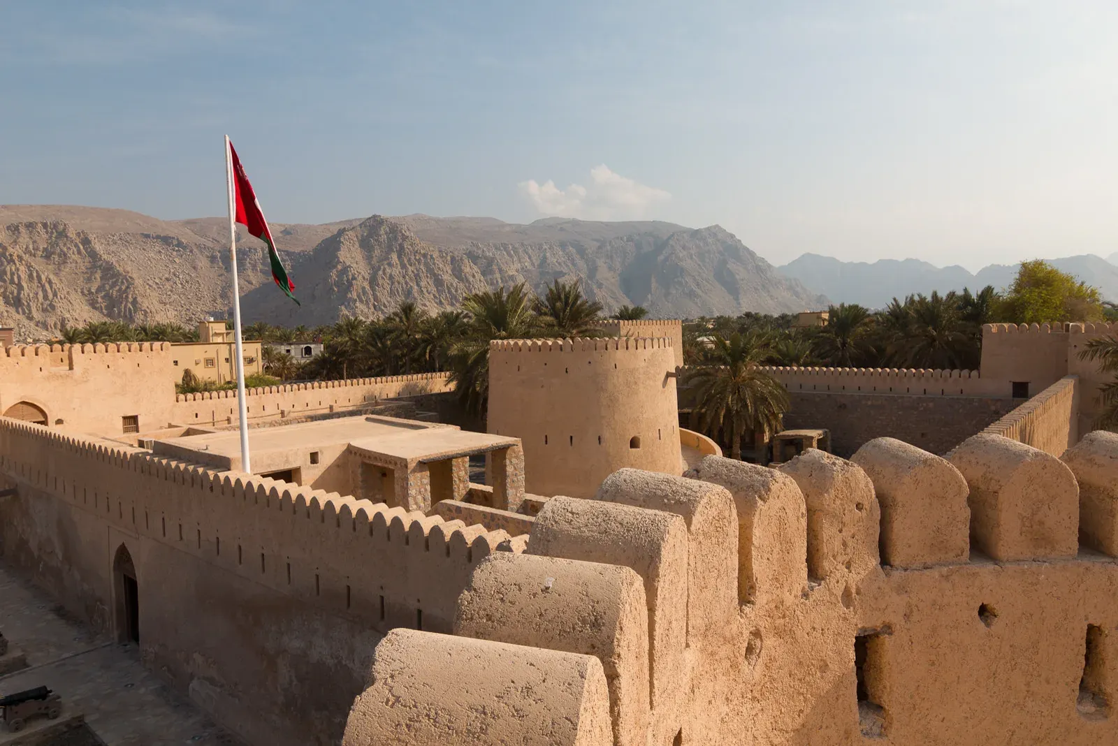 De kantelen van Khasab Fort met de Omaanse vlag, uitkijkend over de palmtuinen en de kale bergen van Musandam