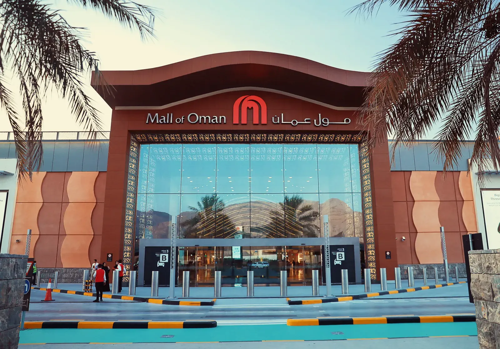 De moderne entree van de Mall of Oman met het logo boven de glazen pui, gezien tussen de palmen door bij avondlicht