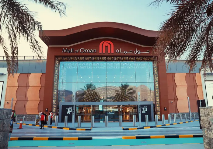 De moderne entree van de Mall of Oman met het logo boven de glazen pui, gezien tussen de palmen door bij avondlicht