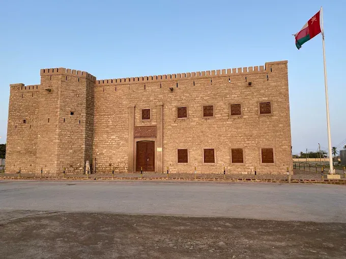 De voorgevel van Mirbat Castle in zandkleurige steen met kantelen, wachttoren en de Omaanse vlag bij avondlicht