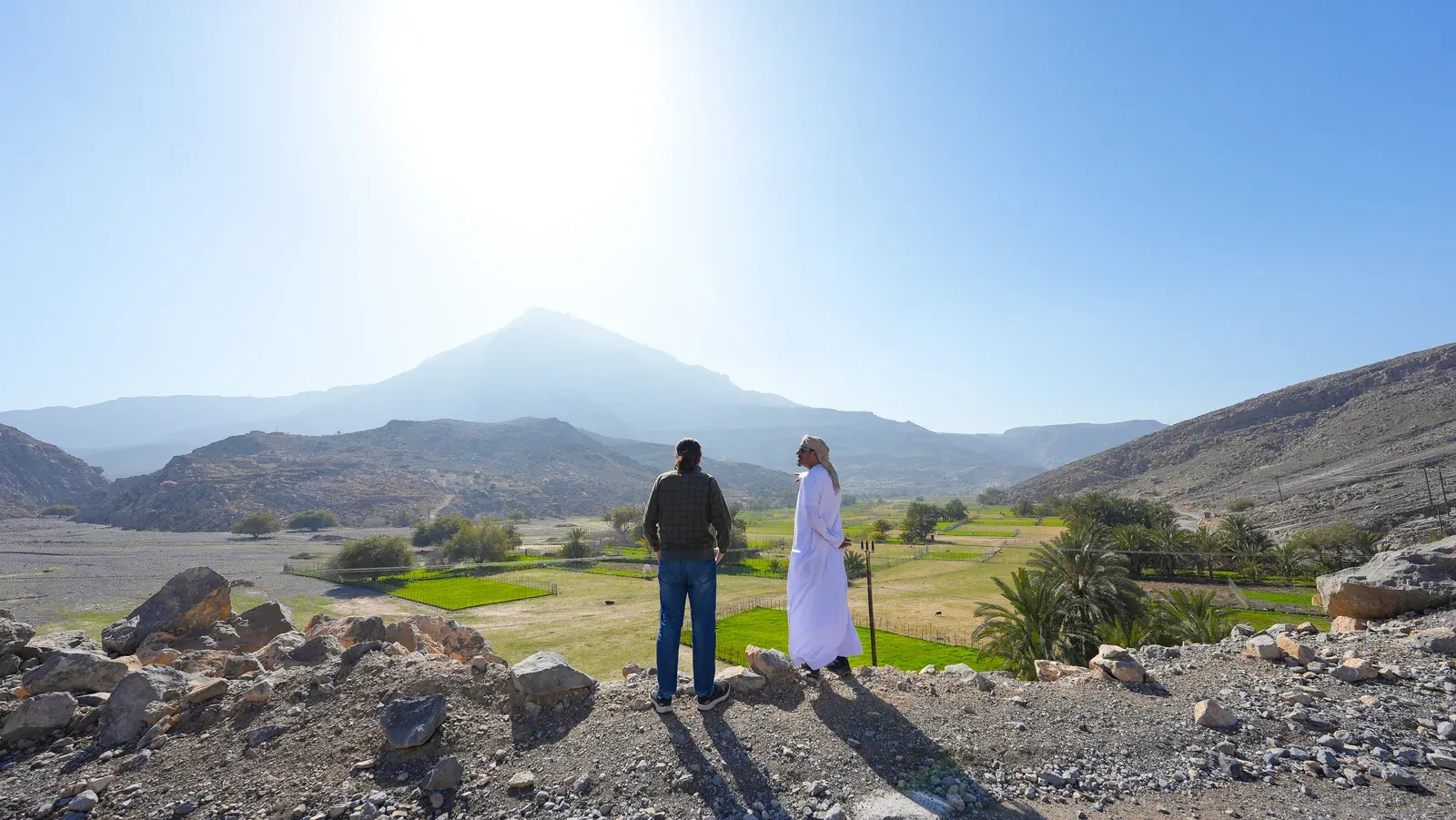 Een gids en reiziger kijken uit over de groene akkers van het Sayh-plateau, met de bergtop van Jebel Harim in de verte