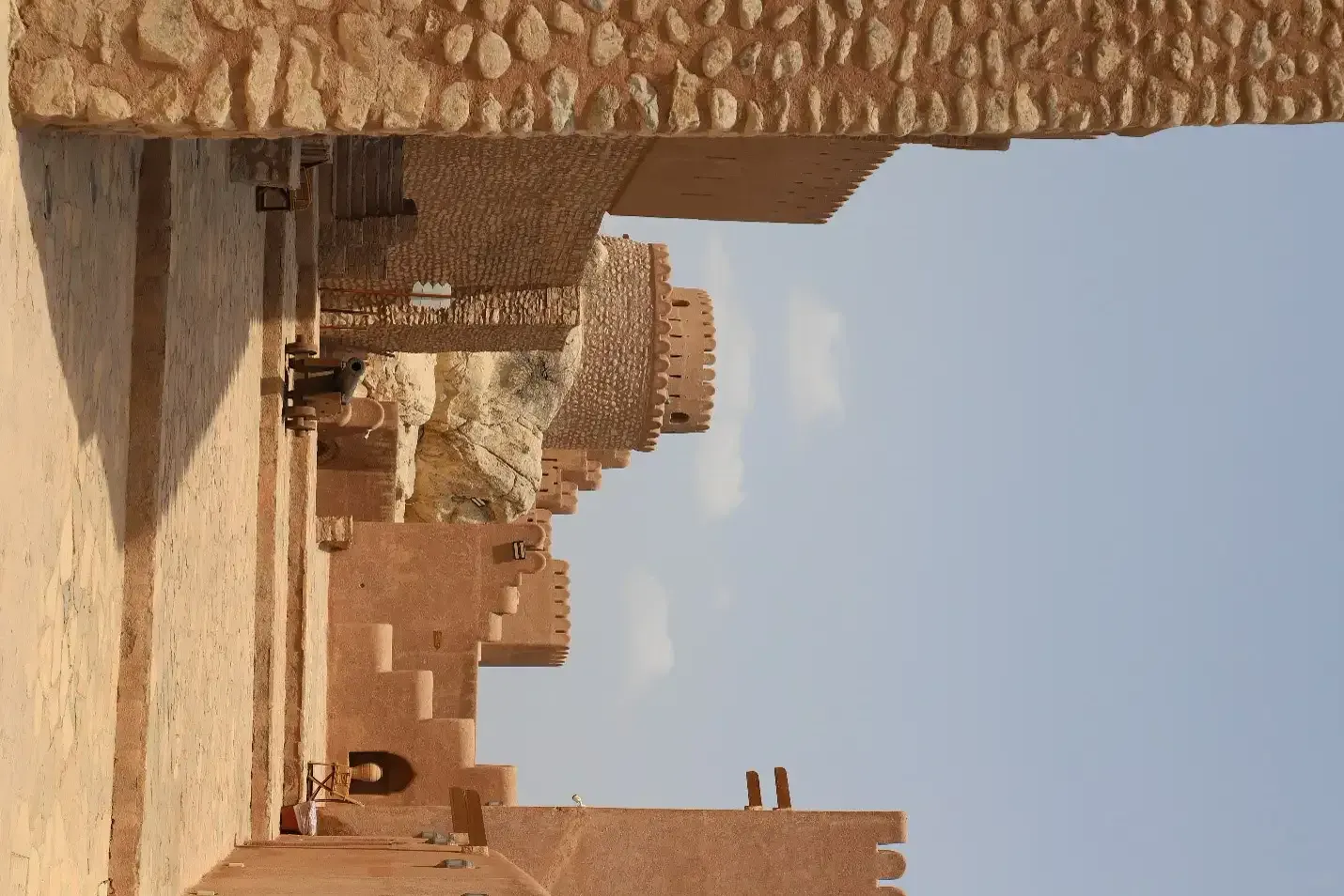De geplaveide binnenplaats van het Nakhl-fort met een oud kanon, ronde wachttoren en lemen muren tegen een blauwe lucht