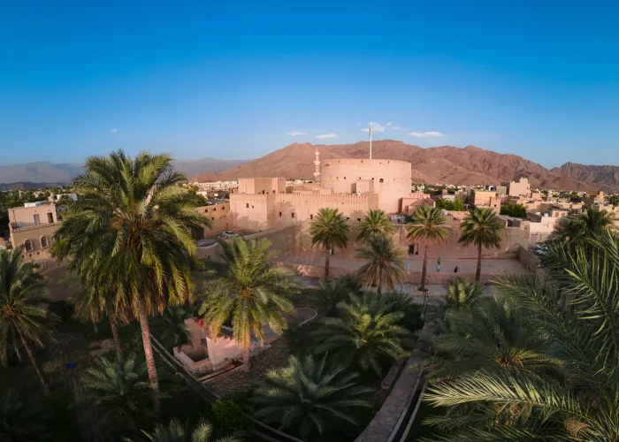 Luchtfoto van Nizwa Fort met zijn karakteristieke ronde toren, omringd door palmtuinen met de Hajar-bergen op de achtergrond