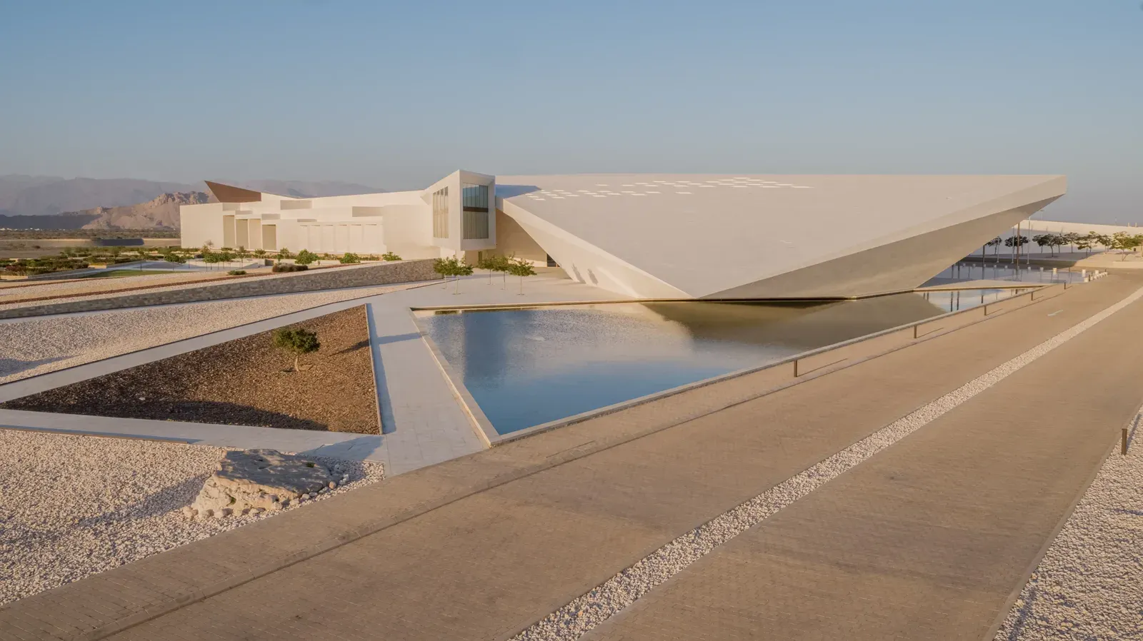 Het scherp toelopende, moderne exterieur van het Oman Across Ages Museum weerspiegeld in een lange waterpartij