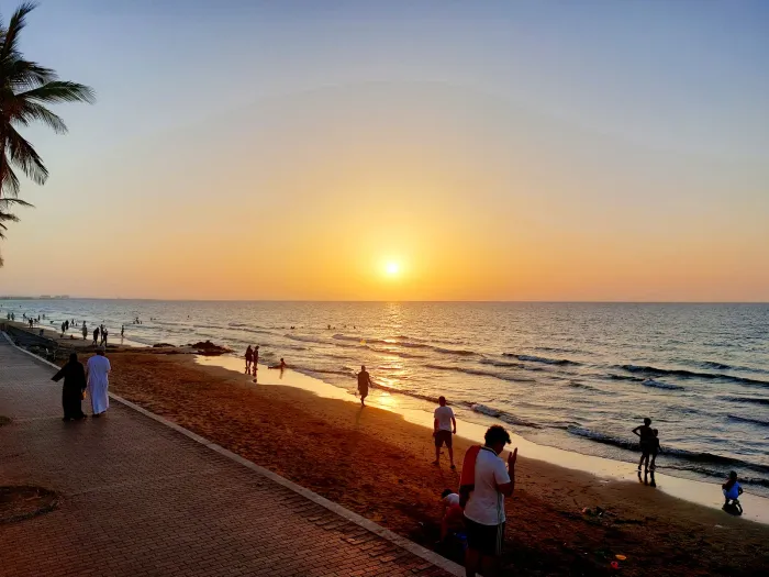 Gouden zonsondergang boven de zee bij Qurum Beach, met wandelaars als silhouet op de brede boulevard