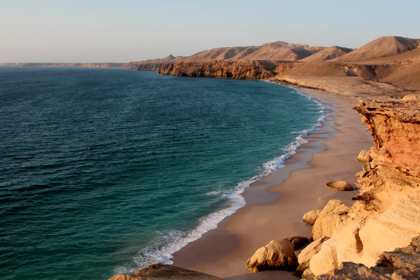 Een verlaten zandstrand met turkooizen branding en rossige kliffen die in de verte uitlopen langs de kust van Ras Al Jinz