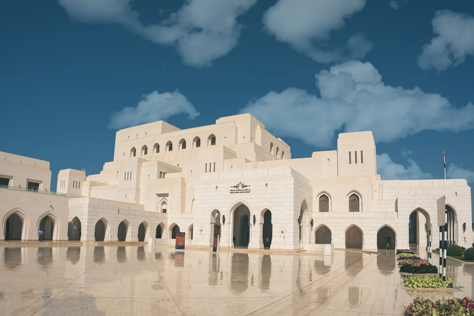 Het Royal Opera House Muscat in wit travertijn, weerspiegeld in het natte plein onder een blauwe lucht