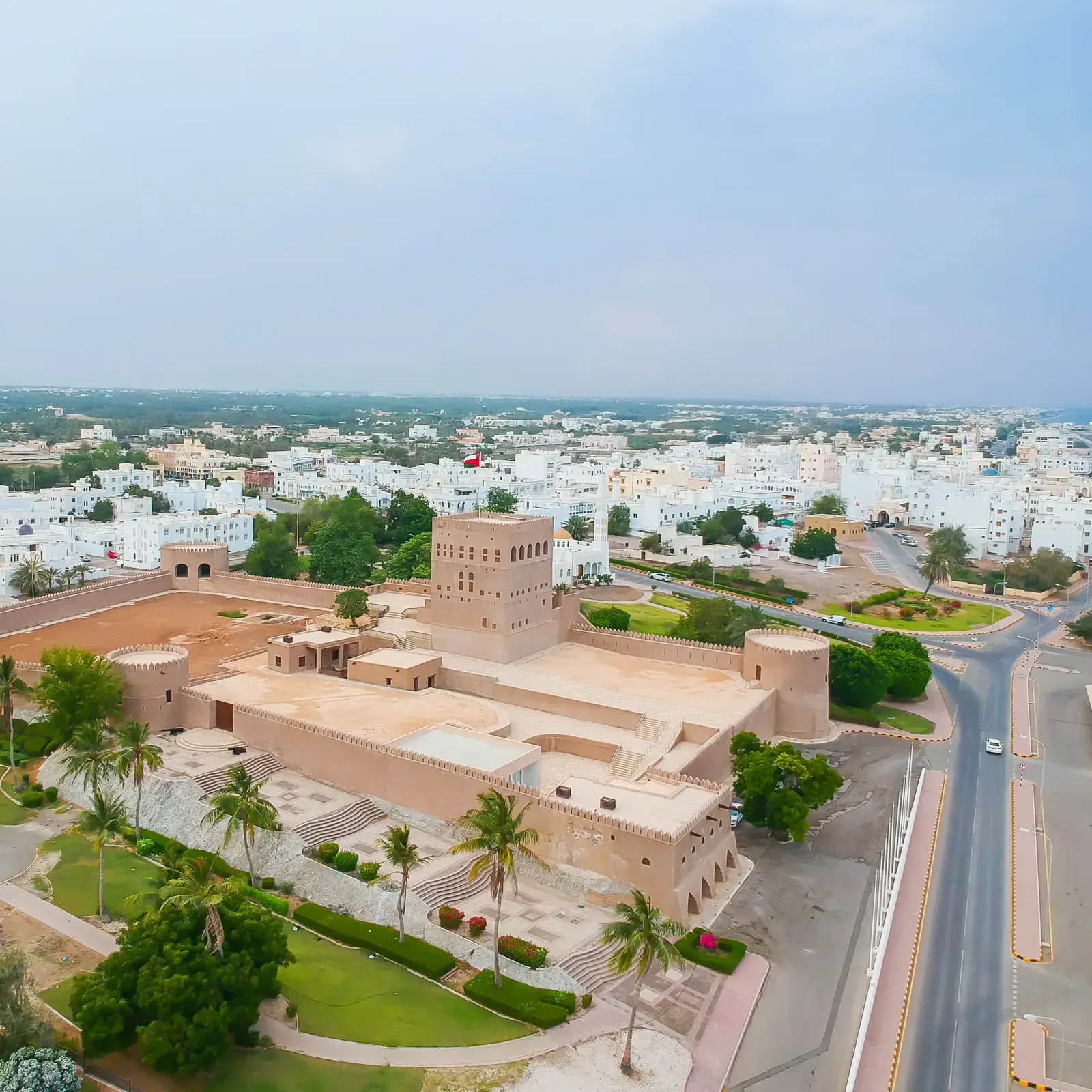 Luchtfoto van het witgekalkte Sohar Fort met torens, groene tuinen en palmen midden in de stad Sohar