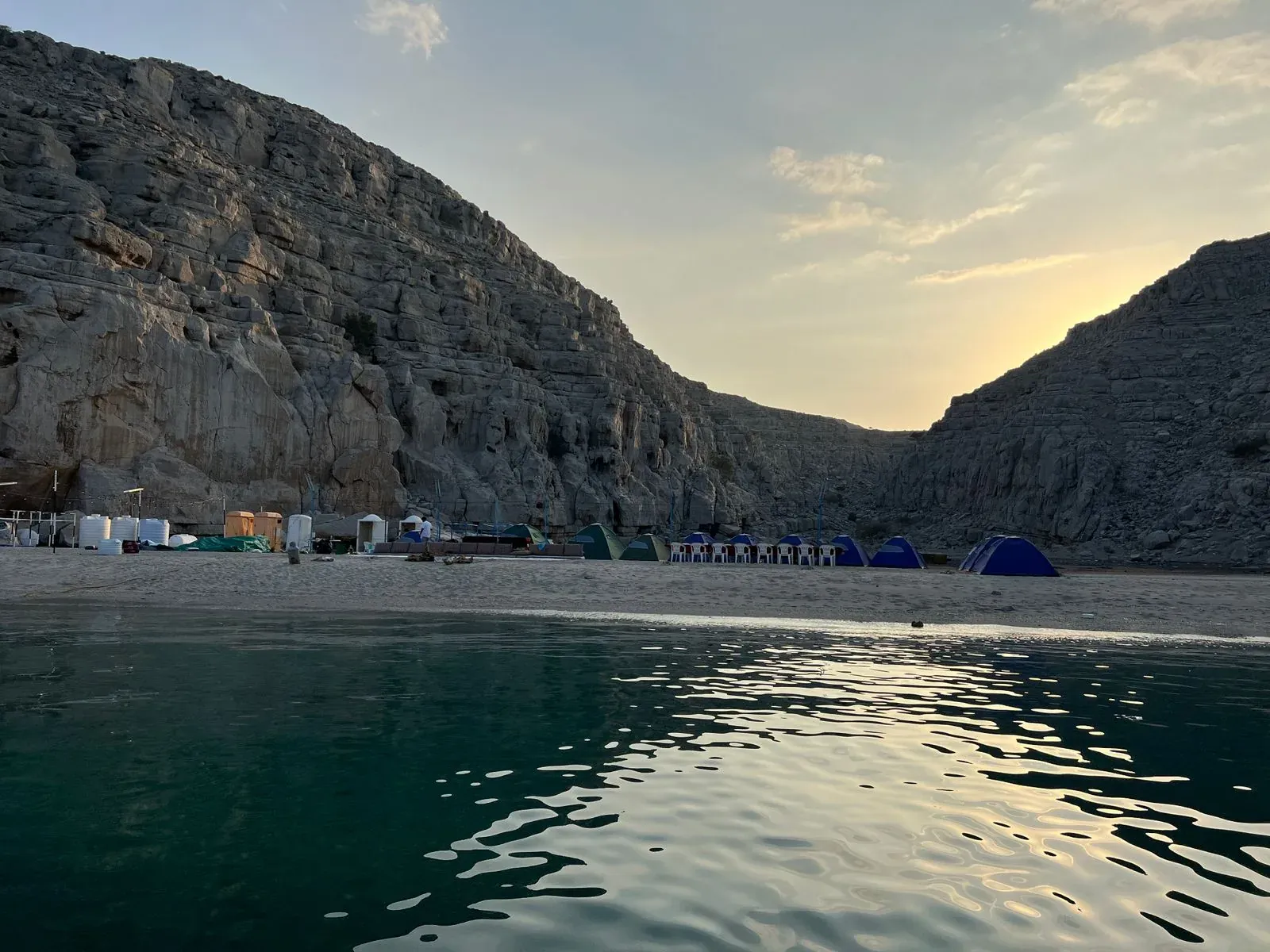Een afgelegen kiezelstrand tussen de fjordwanden van Musandam bij schemering, met kampeertenten aan het water