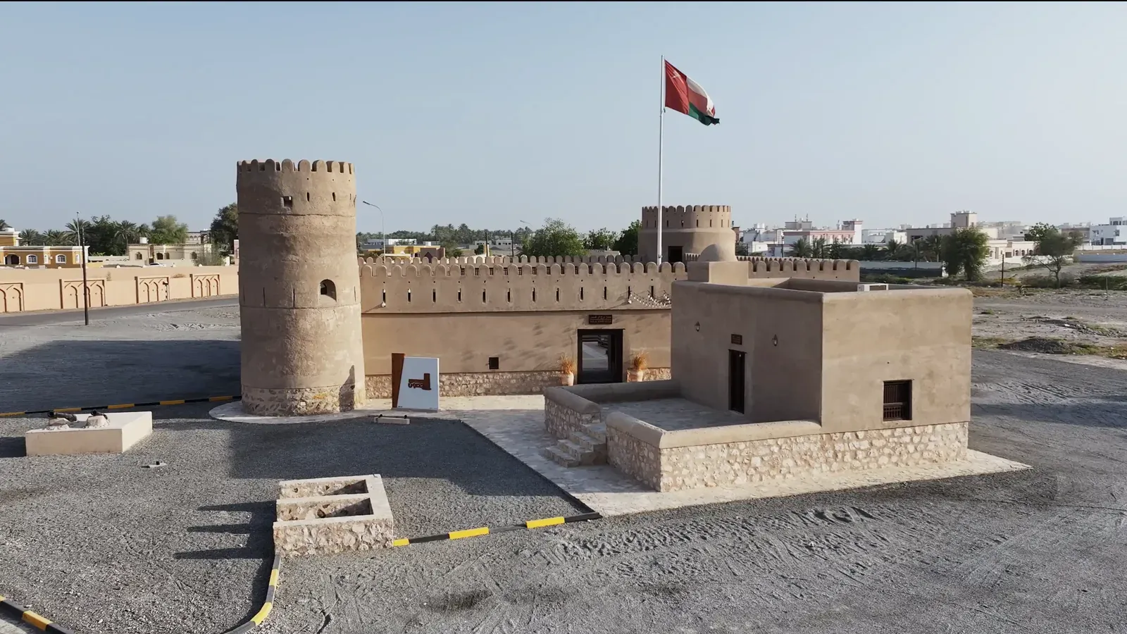 Luchtfoto van het gerestaureerde fort Sur Burj al Khamis met ronde toren, vestingmuur en de Omaanse vlag onder een heldere lucht