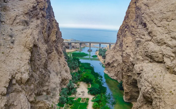 Blik door de steile kalkstenen rotswanden van Wadi Shab op de groene oase, een brug en de zee in de verte